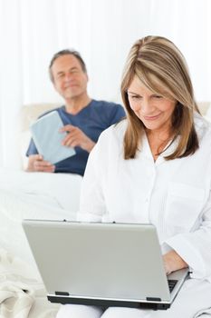 Woman looking at her laptop while her husband is reading on the bed