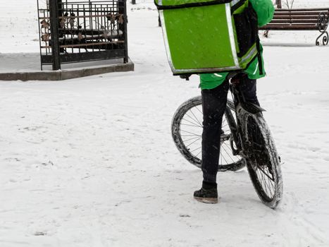 24-hour delivery service from cafes and restaurants. Take-away courier on a bike with a green isothermal backpack rides fast. Courier delivers food by bicycle in winter to avoid traffic jams