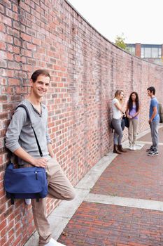 Portrait of a male student leaning on a wall while his friends are talking outside a building
