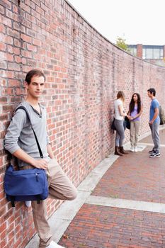 Portrait of a student leaning on a wall while his friends are talking outside a building