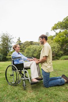 Blonde woman in wheelchair with partner kneeling beside her in the park on sunny day