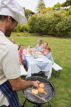 Smiling extended family having a barbecue being cooked by father in chefs hat outside in sunshine