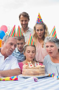 Cheeful family watching girl blowing out candles at birthday party outside at picnic table