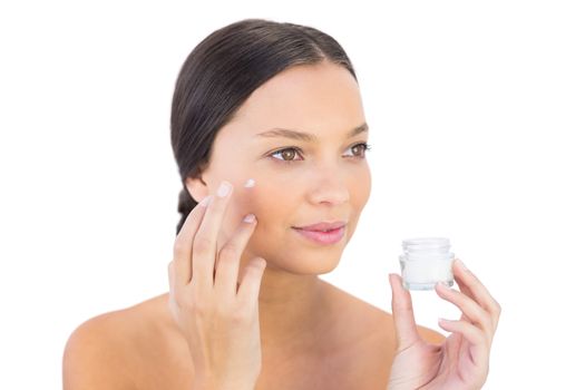 Brunette woman spreading beauty cream on her cheek against white background