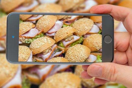 Cold appetizers on tray. hamburger with bacon and herbs in several rows. Sandwiches in smartphone screen.