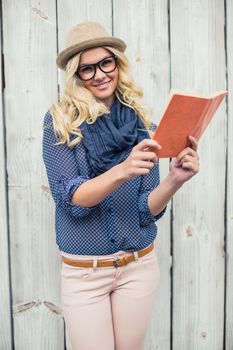 Smiling fashionable blonde holding book outdoors on wooden background