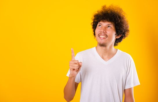 Asian Portrait happy young man curly hair White t-shirt pointing fingers away copy space isolated yellow background