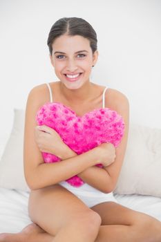 Romantic young brown haired model in white pajamas hugging a heartshaped pillow in bright bedroom