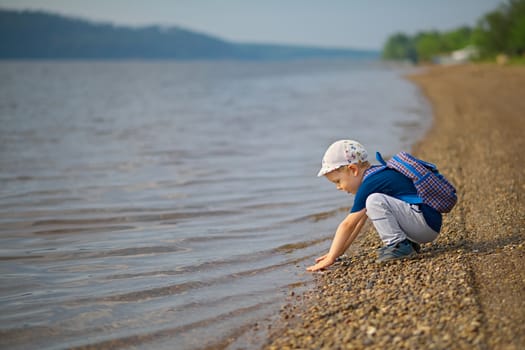 Little, four years old, boy touches the water with his hands in a large river.