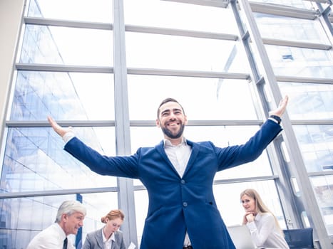 Young joyful businessman standing with arms raised smiling , team of his colleagues sitting at the table in the background