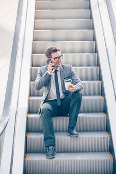 Businessman sitting on escalator talking on phone and holding coffee in hand