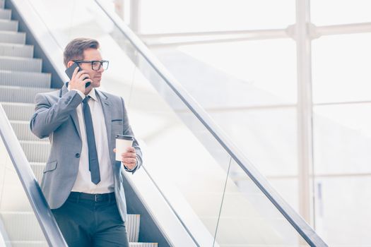 Businessman on escalator talking on phone and holding coffee in hand