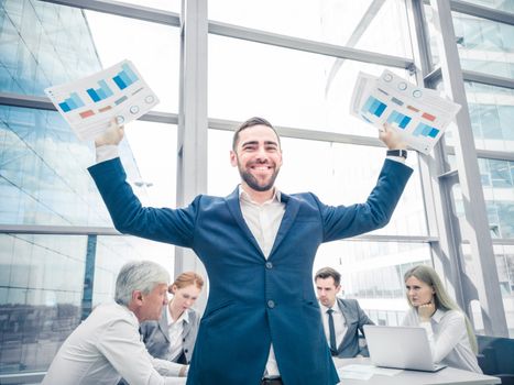 Young joyful businessman standing with arms raised holding data statistics diagrams and smiling , team of his colleagues sitting at the table in the background