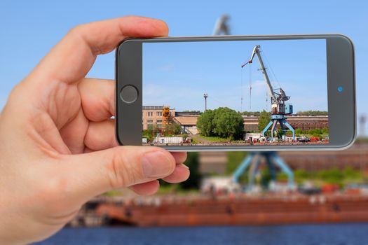 Cranes operate in river port. Cargo port on river. Construction crane in screen of smartphone.