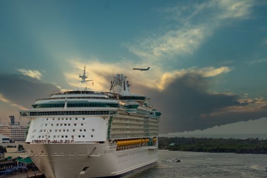 A large cruise ship anchored at a pier with a commercial airplane flying over at dusk