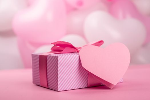 Valentine Day gift in a box wrapped in striped paper and tied with silk ribbon bow and heart shaped greeting card on pink balloons background with copy space for text