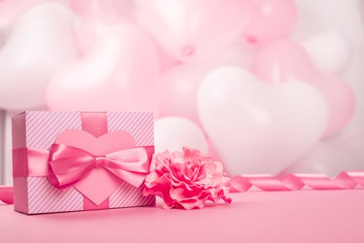 Valentine Day gift in a box wrapped in striped paper and tied with silk ribbon bow and heart shaped greeting card on pink balloons background with copy space for text