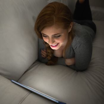 Smiling redhead lying on the couch using her tablet pc at home in the sitting room