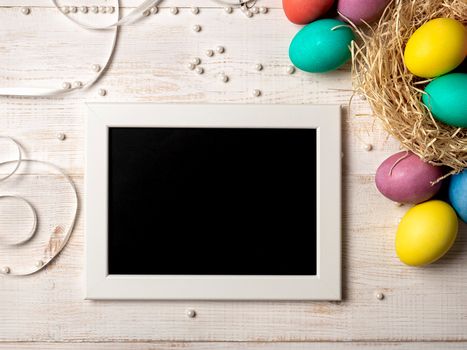 Easter concept. Colorful eggs on white wooden background with empty chalkboard. Copy space for greetings, text or design. Top down view or flat lay