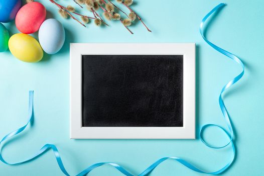 Orthodox Easter concept. Colorful eggs and pussy willow branches on blue background with empty chalkboard. Copy space for greetings, text or design. Top down view or flat lay