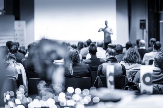 Male public speaker giving a talk at business meeting conference. Rear view of unrecognized people sitting in audience in the conference hall. Business and entrepreneurship concept. Blue toned.