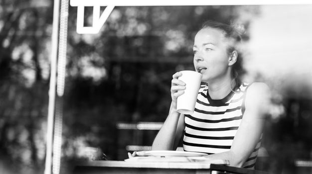 Young caucasian woman sitting alone in coffee shop drinking american coffee, people watching, thoughtfully looking trough the coffee shop window. Black and white image.