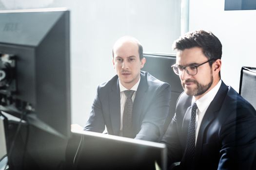 Image of two thoughtful businessmen looking at data on multiple computer screens, solving business issue at business meeting in modern corporate office. Business success concept.