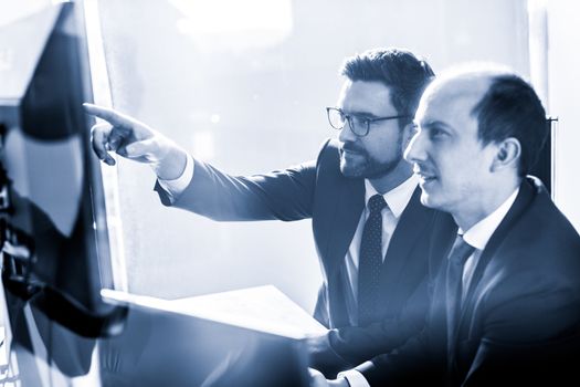 Image of two thoughtful businessmen looking at data on multiple computer screens, solving business issue at business meeting in modern corporate office. Business success concept. Blue toned.