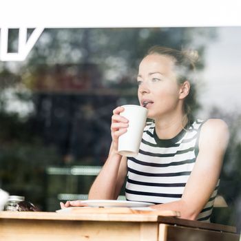 Young caucasian woman sitting alone in coffee shop drinking american coffee, people watching, thoughtfully looking trough the coffee shop window.