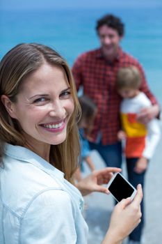 Portrait of cheerful mother holding cellphone while family in background at beach