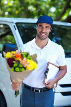 Portrait of happy delivery man holding flower bouquet and clipboard while standing by van