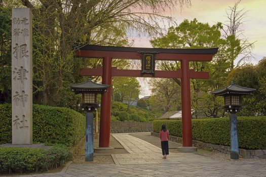 Woman standing at the entrance of the Shintoist Nezu shrine of the 18th century in Tokyo with its big red Torii portal and lanterns at sunset.