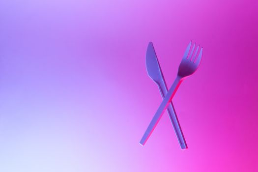 Disposable appliances plastic white fork and knife levitate in neon light, pink blue background, copy space. Eco, zero waste concept. Front view. Horizontal.