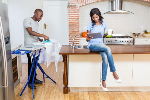 Young woman holding coffee mug and using digital tablet while man ironing clothes in the background