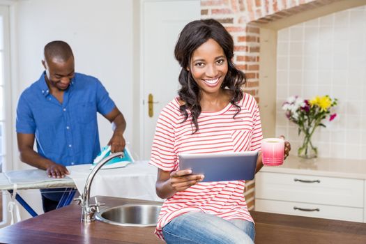 Young woman holding coffee mug and using digital tablet while man ironing clothes in the background