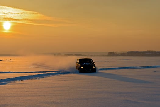 The car rides on the ice of a frozen river in the snow in the rays of the setting sun.