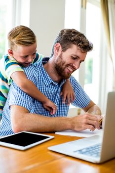 Smiling son leaning on father working with laptop and tablet at home 
