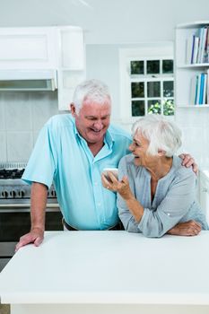 Smiling senior woman showing phone to man while standing in kitchen