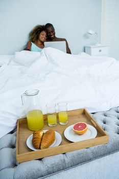 Young couple using a laptop with breakfast tray ready on bed in bedroom