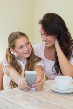 Cute little girl with mother photographing through mobile phone at table in house