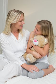 Happy girl embracing teddy bear while looking at mother in bed at home