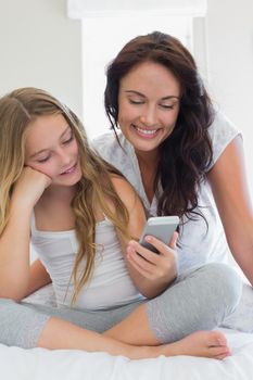 Mother and daughter using mobile phone together while sitting in bed at home