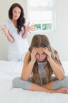 Irritated girl sitting in bed while mother scolding her in background at home