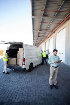 View of manager is using a tablet in front of workers are loading a truck in front of a warehouse