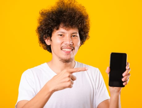 Happy asian handsome young man with curly hair holding blank screen smart phone and pointing finger isolate on yellow background
