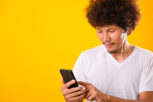 Happy asian handsome man with curly hair he smiling enjoying listening to music on earphones using a mobile smartphone isolate on yellow background