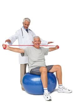 Male physiotherapist looking at senior man sit on exercise ball with yoga belt over white background