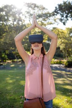 Woman standing with her hands joint while using a VR headset in the park on a sunny day