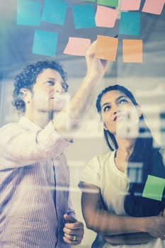 Businessman and businesswoman discussing over adhesive notes on glass wall in office