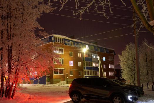 The night street is illuminated by winter lights from the windows of the house and the headlights of cars